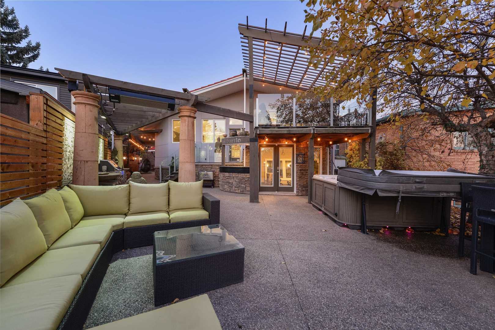 Backyard patio with cement floor; green sectional couch on left, hot tub on right, pergola and bar in background leading to house