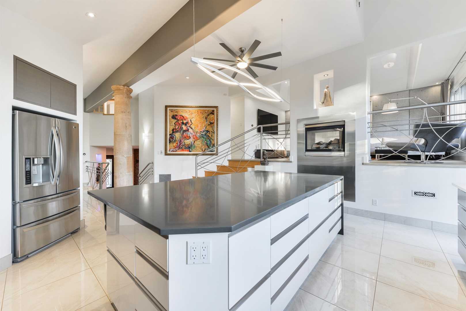 Kitchen with white tile floor, white ceiling and walls; white rectangular island with grey counter underneath tube lighting and silver ceiling fan; two-way fireplace separates kitchen from risen living room