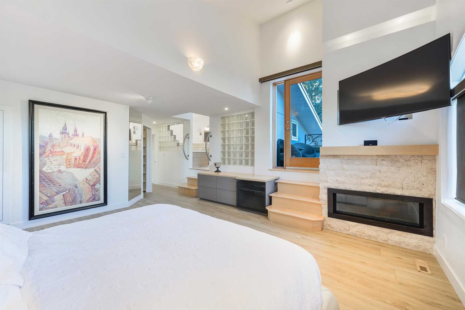 Master bedroom with light hardwood floor, white ceiling and walls; wood stairs leading to patio next to mounted wall TV; stairs to left lead to en suite