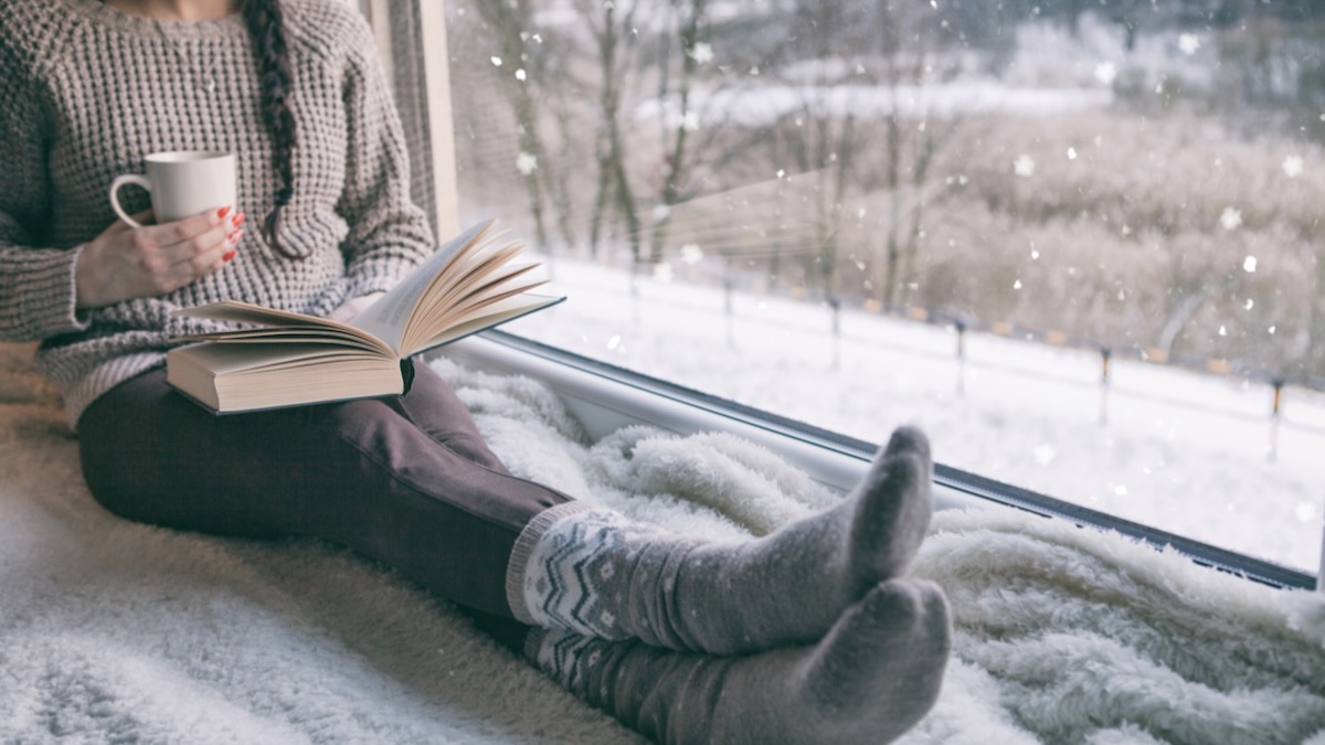 Woman sitting by the window reading book drinking coffee. Winter snowing landscape outside