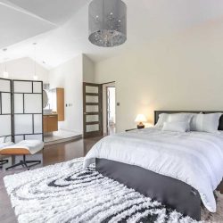Master suite with grey hardwood floor and white walls; a grey circular chandelier hangs mid-room over the grey bed with white duvet to the right, on top of black and white shag carpet; a light wood and white chair and foot stool sit left, in front of white-with-black lines room divider, sectioning off the en suite