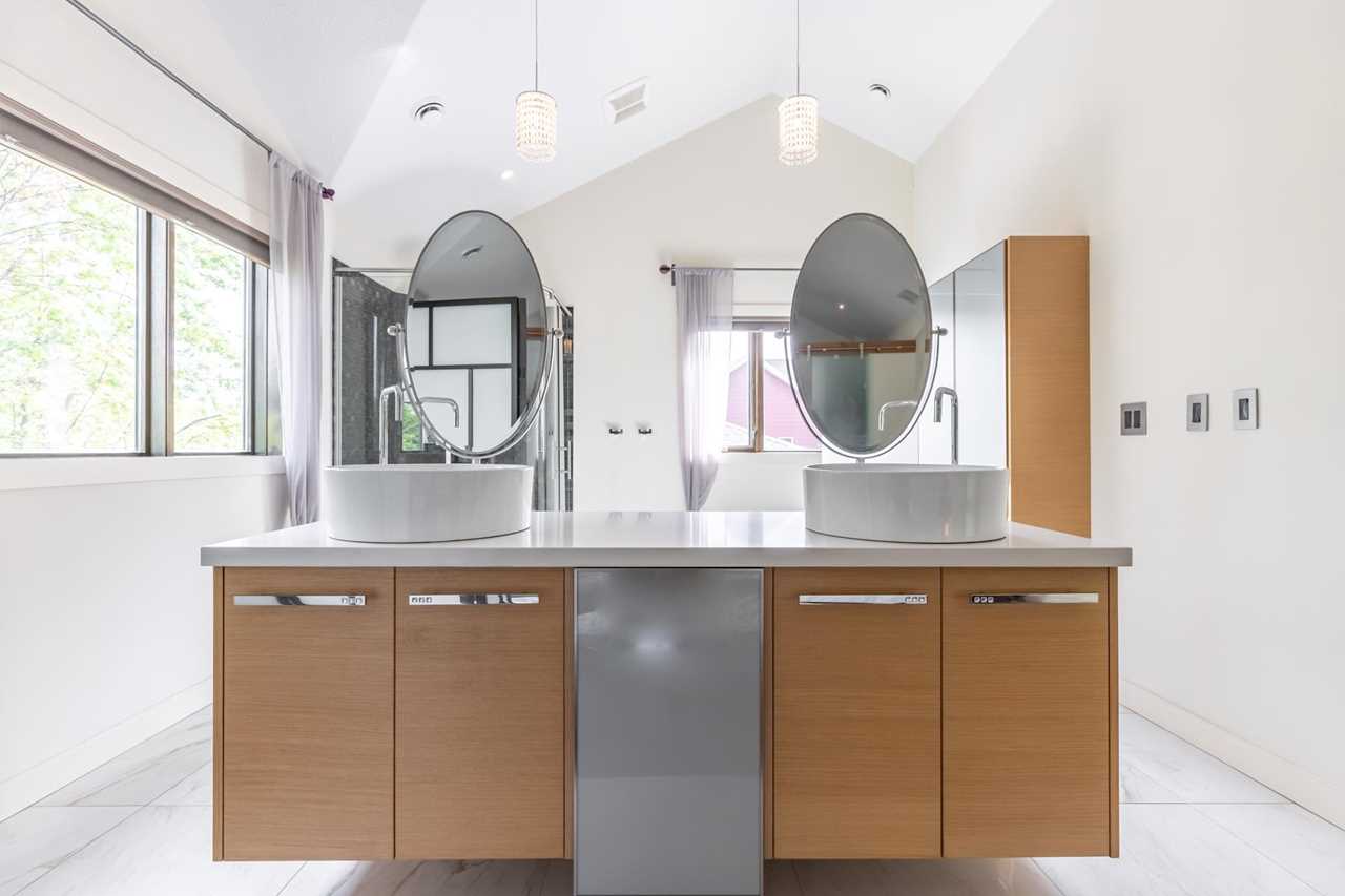 En suite with white tile floor, white ceiling and walls; free standing light brown and grey cabinet unit with built-in sinks and individual oval mirrors above; light brown and silver cabinet back and to the right; windows in background and on the left; two white lights hang above