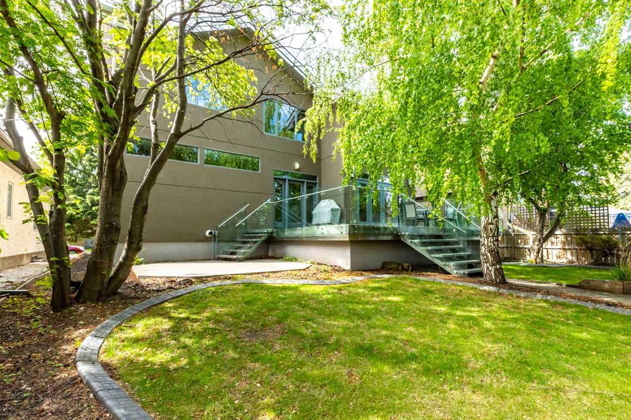 Backyard with green grass; two mature trees on right and left; glass-railed patio with steps on right and left in front of the home's beige back wall 