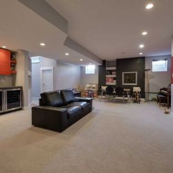 Basement, white ceiling, walls and floor; black couch in front of bar with red cupboard facing wall-mounted TV; children's play area in background
