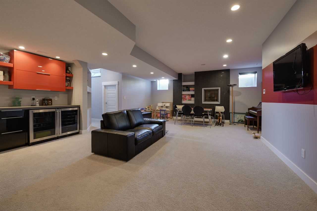 Basement, white ceiling, walls and floor; black couch in front of bar with red cupboard facing wall-mounted TV; children's play area in background