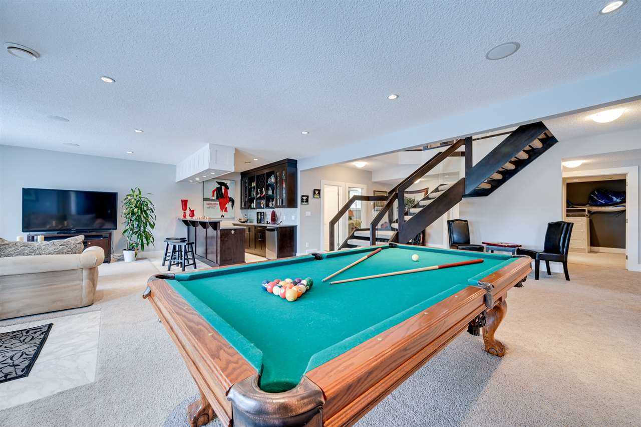 Basement with white walls and light carpet; green-surface pool table in foreground; dark-cabinet bar, fake plant, large TV and light beige couch to the left