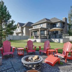 Yard with fire pit surrounded by red Adirondack chairs across the lawn from the house with two-sectioned deck