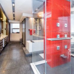 En suite with dark grey, white-lined tile floor; wood cabinetry on left; glass shower with bright red wall and grey tile floor on right, looking out to city view