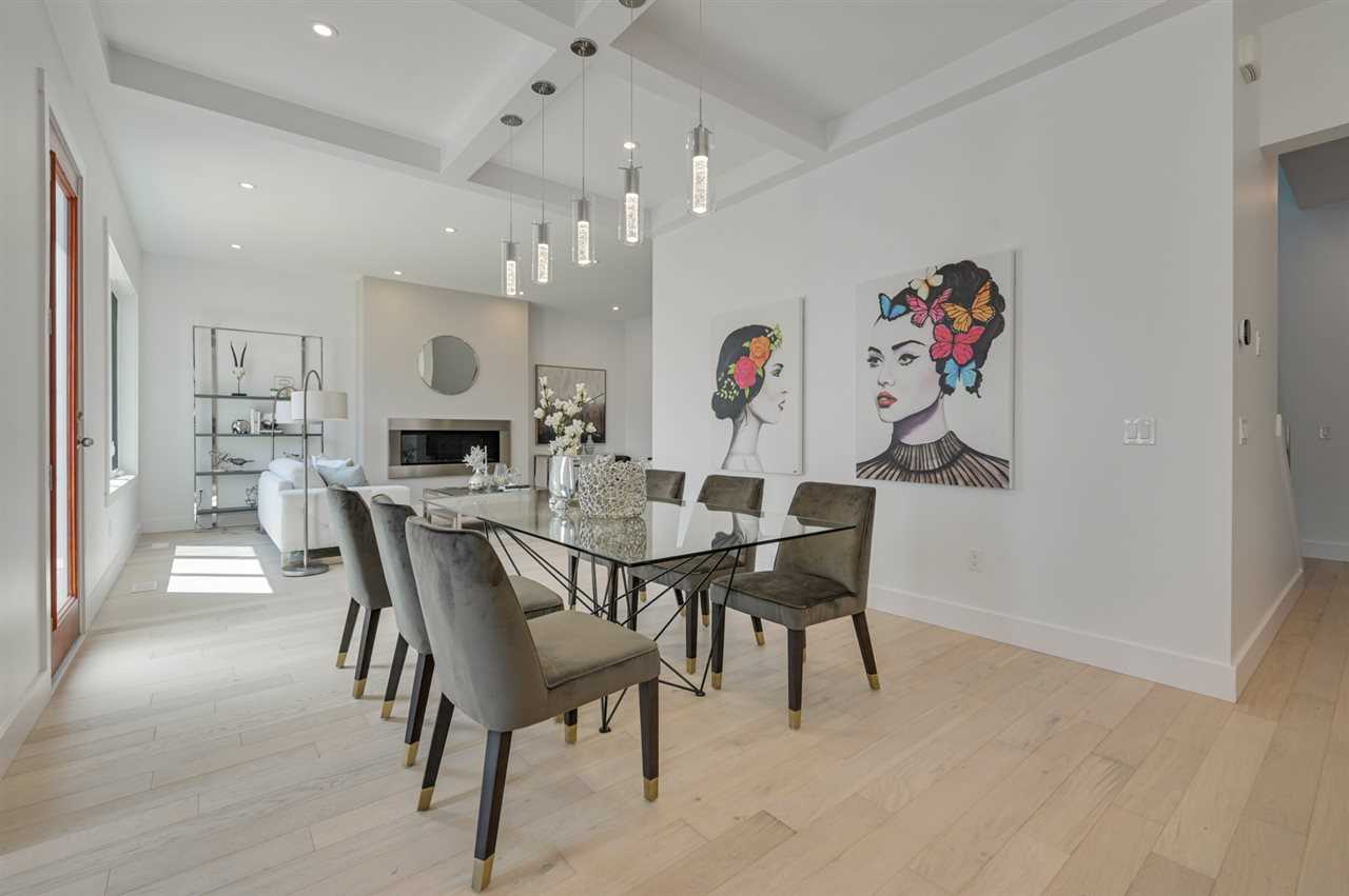 Interior home, dining foreground/living area background; white oak floor, white walls and coffered ceiling with five hanging lights over glass dining table with grey chairs; two black and white paintings of a woman's face with coloured flowers and butterflies in her hair