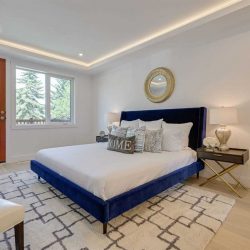 Mater bedroom with white ceiling and walls, white oak floor; track lighting around ceiling perimeter; blue bed frame with white sheets; wood-framed glass door leading to balcony