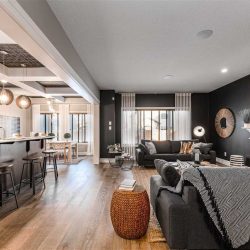 Interior living room/kitchen; hardwood floor, white ceiling; black living room wall with dark grey couches, white fireplace mantle; white ceiling beam separating living room and kitchen; dark wood kitchen island with three stools