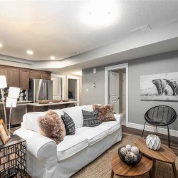 Basement suite with white ceiling and walls, hardwood floor; white couch, small wood coffee tables; wood cupboards in kitchen, triangle pattern backsplash