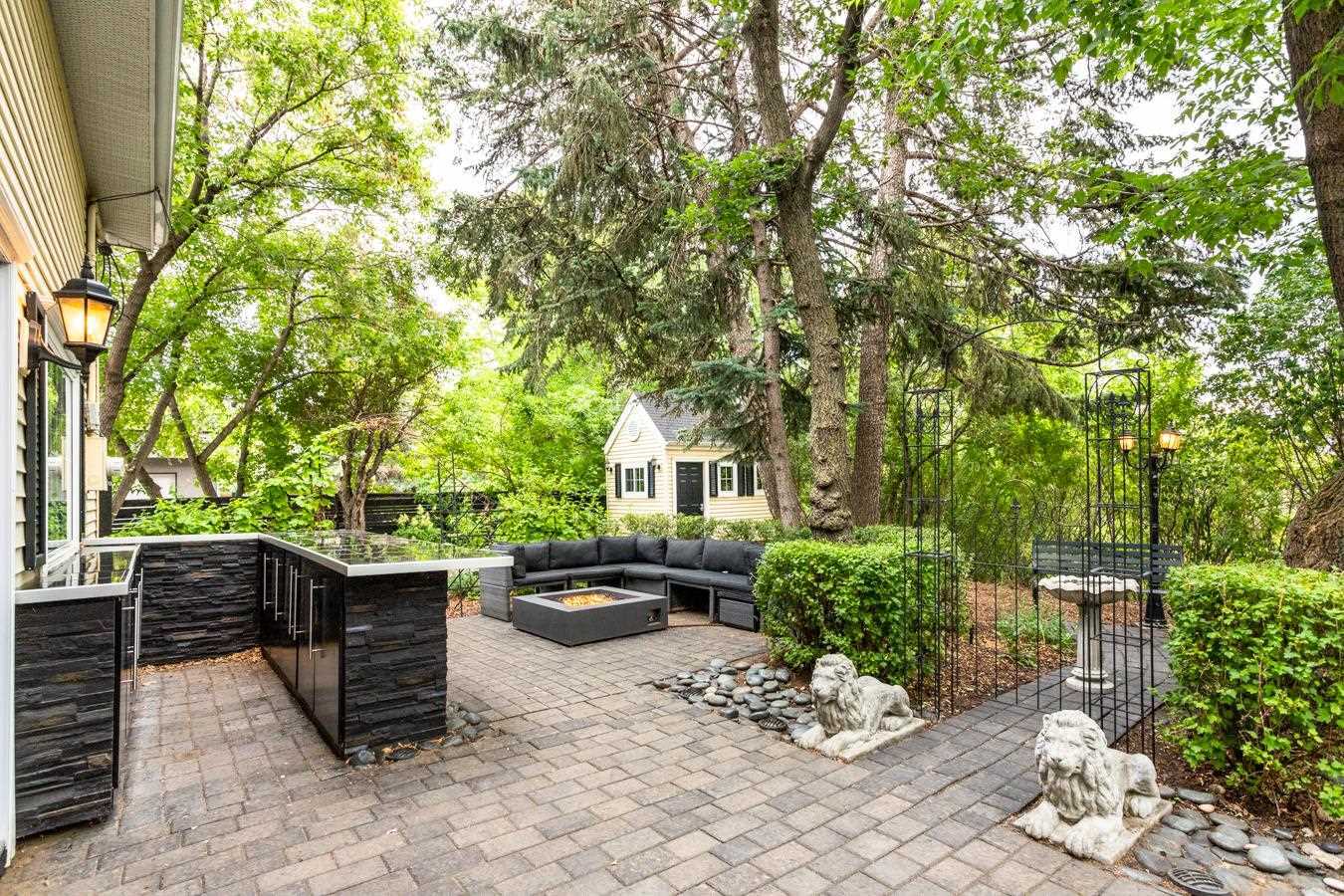 Backyard ground floor patio made of grey stone; wrought iron gate to right, black stone outdoor kitchen to left, black sectional patio couch with square fire pit in middle; shed that looks like a tiny guest house in background