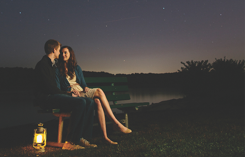 A couple sits by a storm water pond in the evening, with the clear night sky, stars, and a lantern by their side.