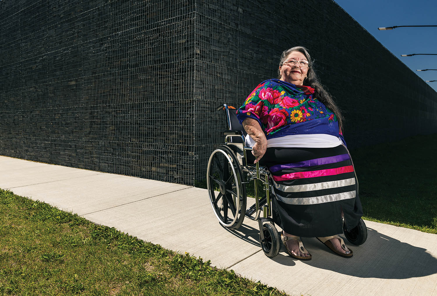 ElderMarggoPariseau_InWheelChaironSidewalk_CementWall_ColourfulScarf_RibbonSkirt