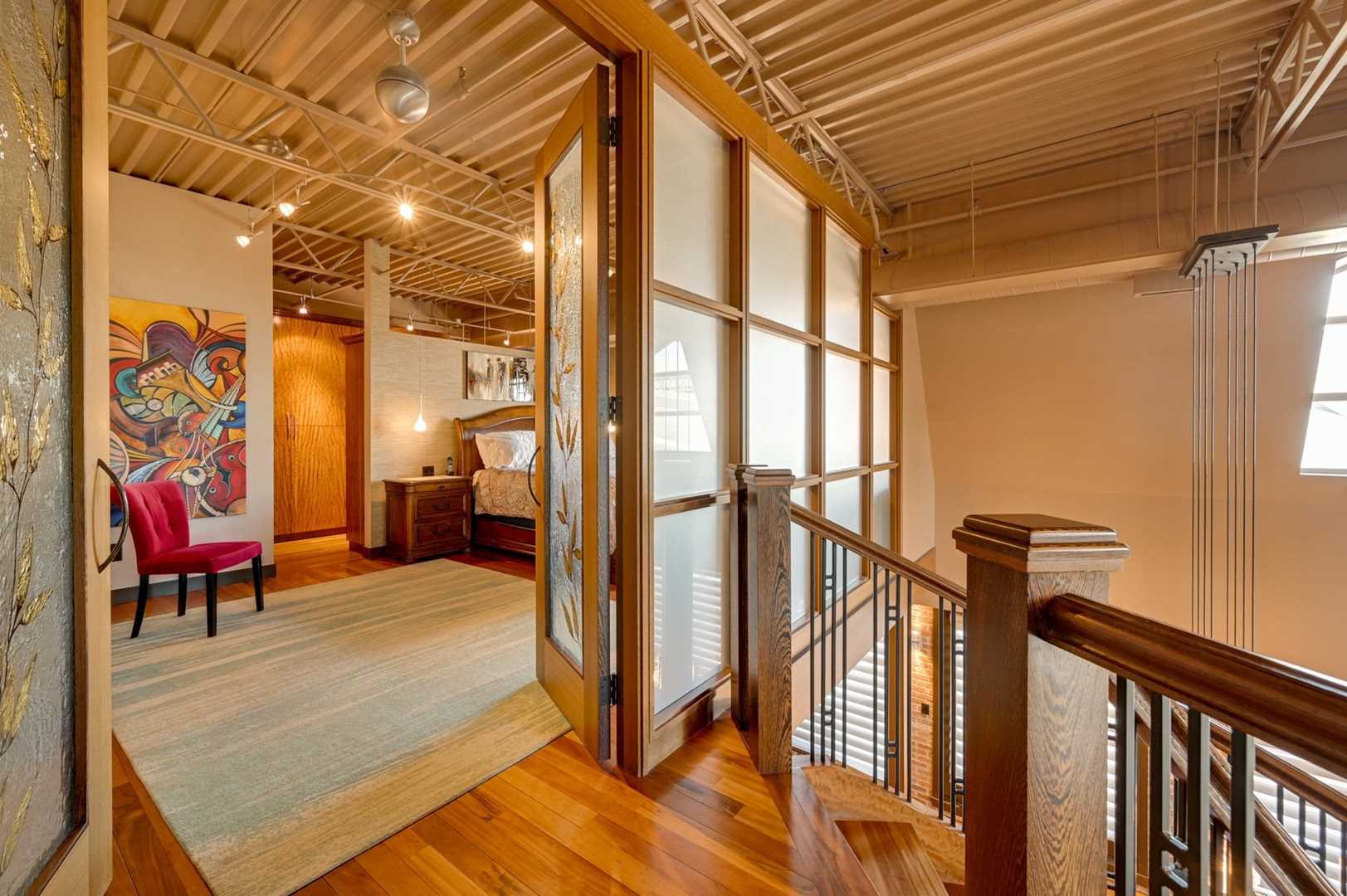 Interior entrance to loft bedroom with tiger wood floor, beige walls and open ceiling; glass doors open to bedroom on left; top of staircase on right