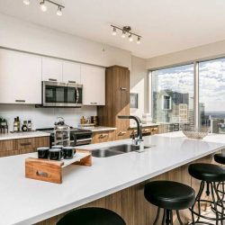 Interior condo kitchen with white marble island countertop with a sink, wood panel cupboards, light wood floor, four black stools; two-storey windows on right