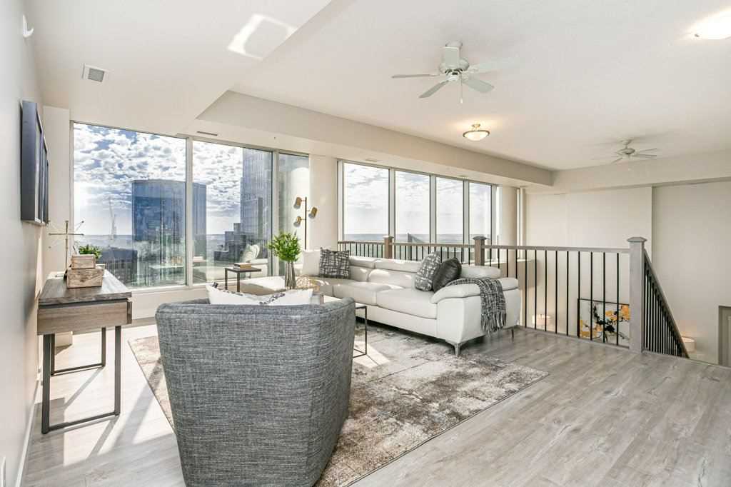 Condo upstairs loft with white ceiling, ceiling fan and walls; light grey hardwood floor; dark grey chair in foreground; white couch along railing in background; large windows behind