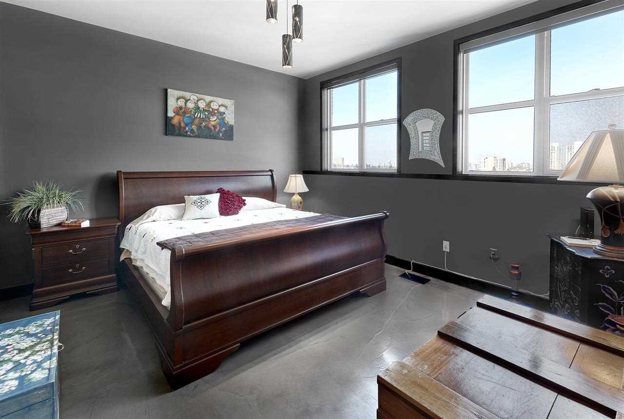 Master bedroom with grey floor and walls, white ceiling; wine-red sleigh bed with white bedding; wine-red nightstand to the left; wood crate on right; two windows on far wall