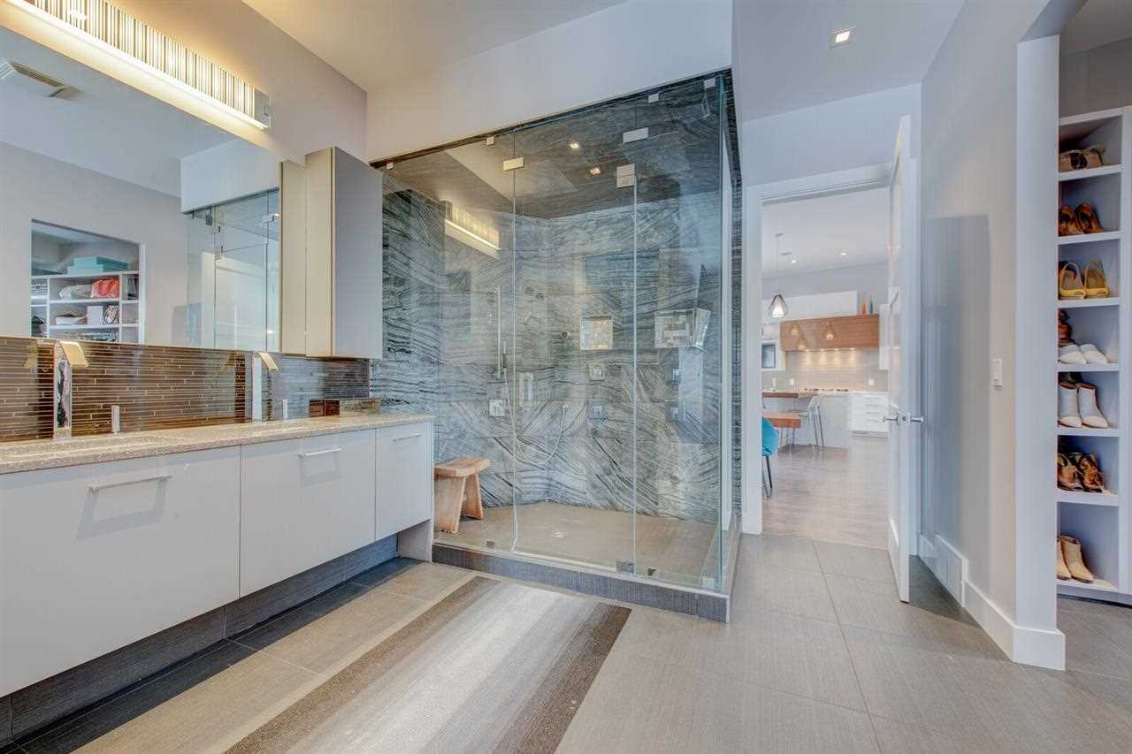 En suite bathroom with vinyl floor, white cabinets, ceiling and walls; glass shower next between open door and two sinks in front of large mirror (on left); on right, vertical shoe storage