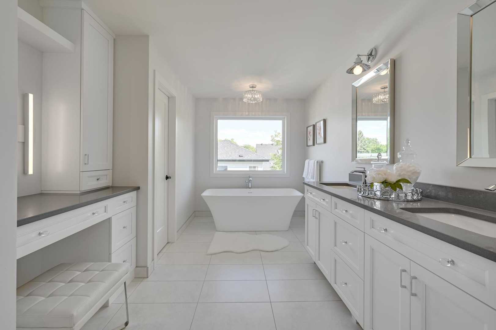 En suite bathroom, white ceiling cupboards and walls; soaker tub in front of window; makeup table and vanity mirror to left