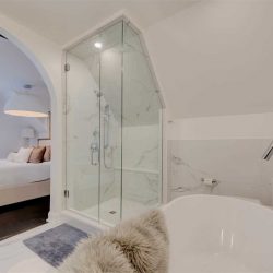 En suite bathroom, white ceiling and walls; white soaker tub with fur blanket overhanging on right; clear stand-up shower slanting with the ceiling in middle; bedroom through the door on left