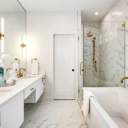 All white en suite with gold finishes and fixtures; tub on right, shower behind it; sliding door to toilet in the middle; double sinks in front of large mirror on left