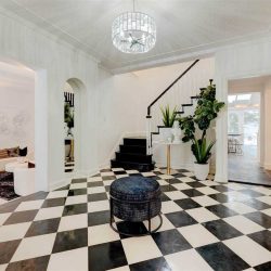 Entrance with black and white checkered floor, single black stool in middle underneath chandelier; white ceiling and walls