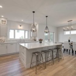 Interior kitchen, light hardwood floor, white ceiling and walls; four steel stools at white waterfall island, two light hanging overhead; white cupboards and pantry to left