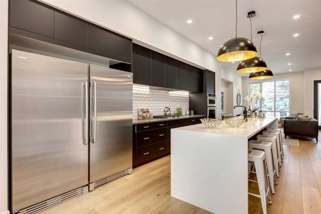 Kitchen with light hardwood floor, white ceiling; white marble waterfall island with five white stools on right, three steel dome lights above; massive fridge on left, black cabinets with white penny-tile backsplash; living room with black couch behind