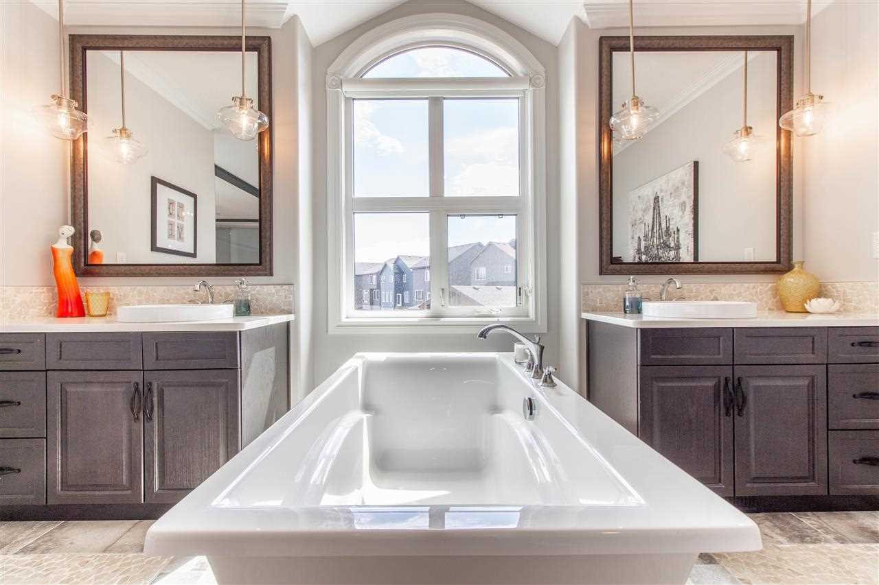 Interior en suite, white soaker tub framed centre with cove-window behind it; a sink on either side, each with a mirror and two hanging lights; dark wood cabinets