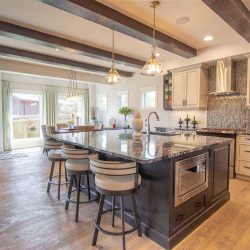 Interior kitchen with light hardwood floor, white cabinets and ceiling with cedar beams; black island with black and white countertop, microwave built into one side, three stools on the other, two hanging lights above; black and white backsplash above stove