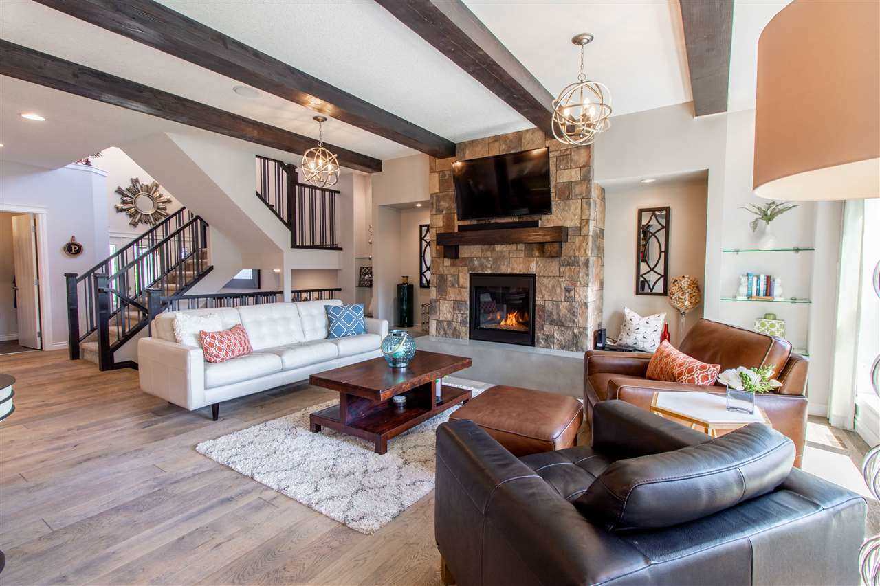 Interior living room with light hardwood floor, white walls and ceiling with decorative cedar beams and two chandeliers; dark and light leather seats on the right, white couch on left, small wood coffee table in between; stone fireplace with TV above in background