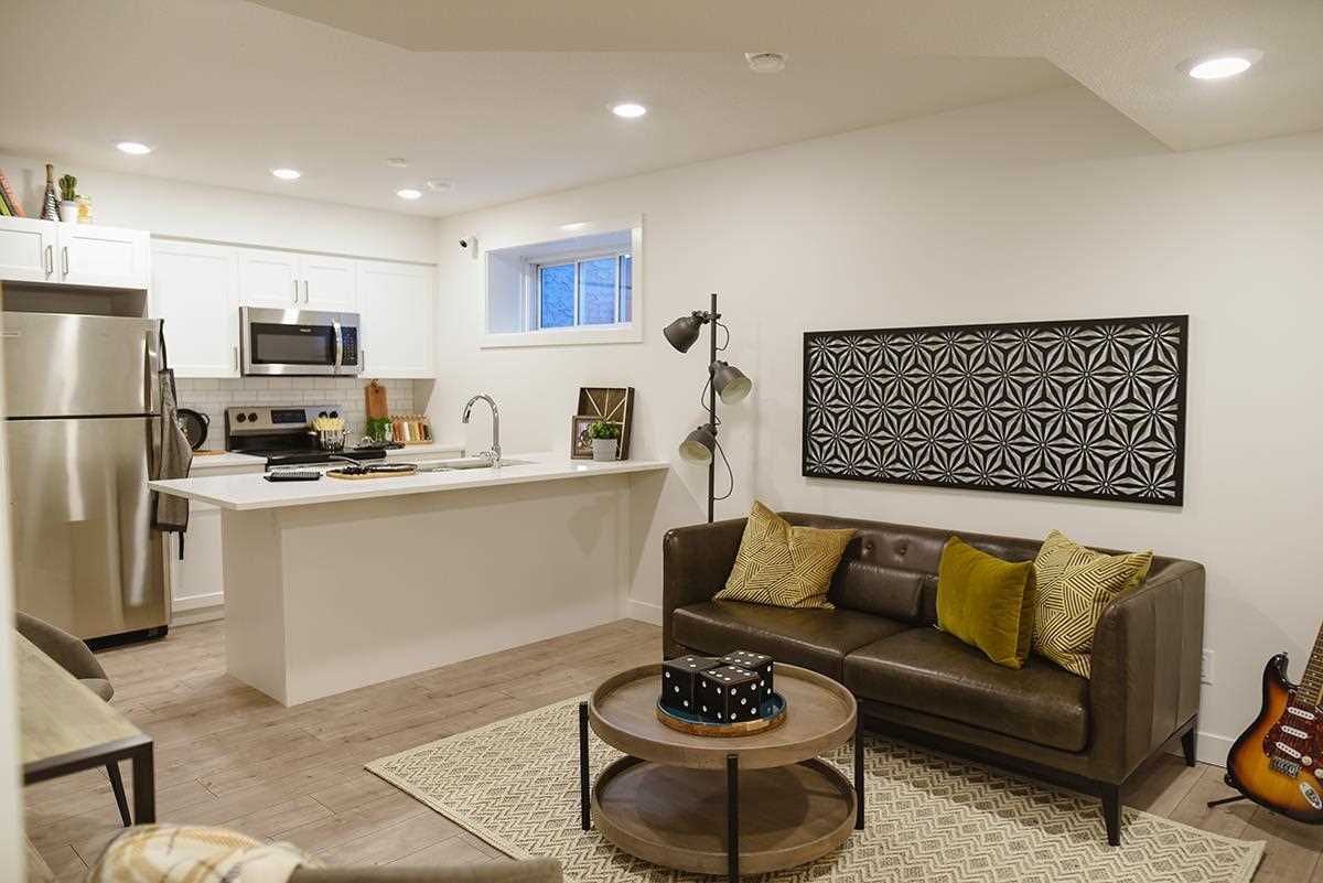 Basement suite with white walls, light grey floor and small window; white kitchen island, brown couch and circular coffee table.