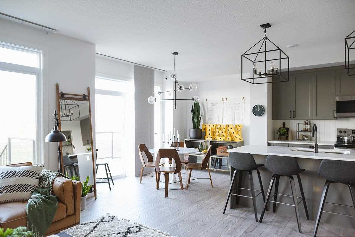 Kitchen, living and dining area; white ceiling and walls, light grey floors, wood chairs at dining table, grey and black stools at kitchen island; Edison light bulb chandeliers. 