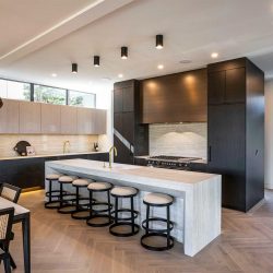 Kitchen with light hardwood floor, white ceiling; white marble waterfall island with six chairs facing black bordered stove area