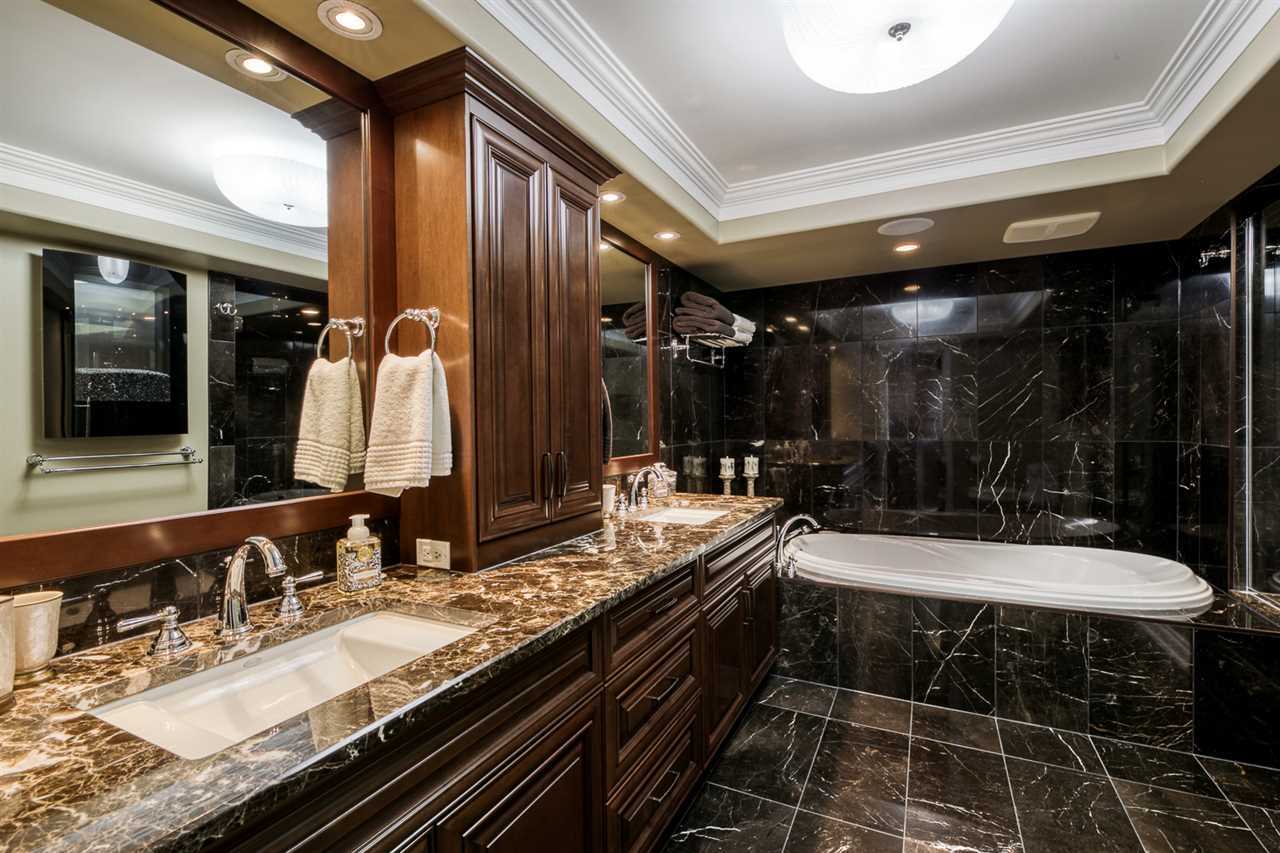 En suite with black tile floor and walls, white ceiling; double sinks in dark marble counter with dark wood cabinet on top and between; dark wood drawers below, on left; white tub in background embedded in the black tile