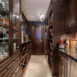 Wine cellar with light tile floor and ceiling, dark wood cabinets on right and left, dark wood door at far end; wine glasses in glass cabinet on left, marble counter with artistic jugs, wine opener and Grand Marnier on right, with mini-fridge below; wine racks beyond on each side