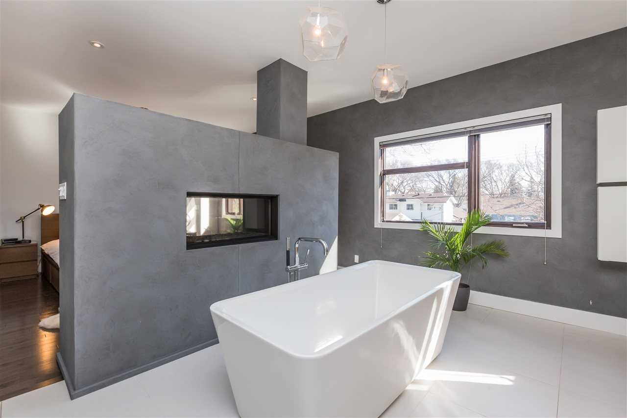 En suite bathroom with white tile floor and grey walls; grey false wall with embedded fireplace separating en suite from bedroom; white soaker tub in the middle, two hanging lights above; large window on back wall with green plant on the floor in front