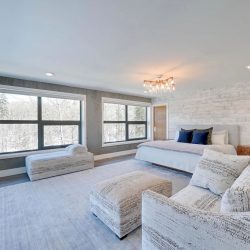 Master bedroom, white ceiling and walls with large windows; light grey bed and couch with chandelier over the bed; wall-mounted TV