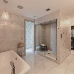 En suite with white tile floor and white marble walls, all speckled with black; freestanding soaker tub on the left, large shower with a black stool on the right; walk-through closet in background