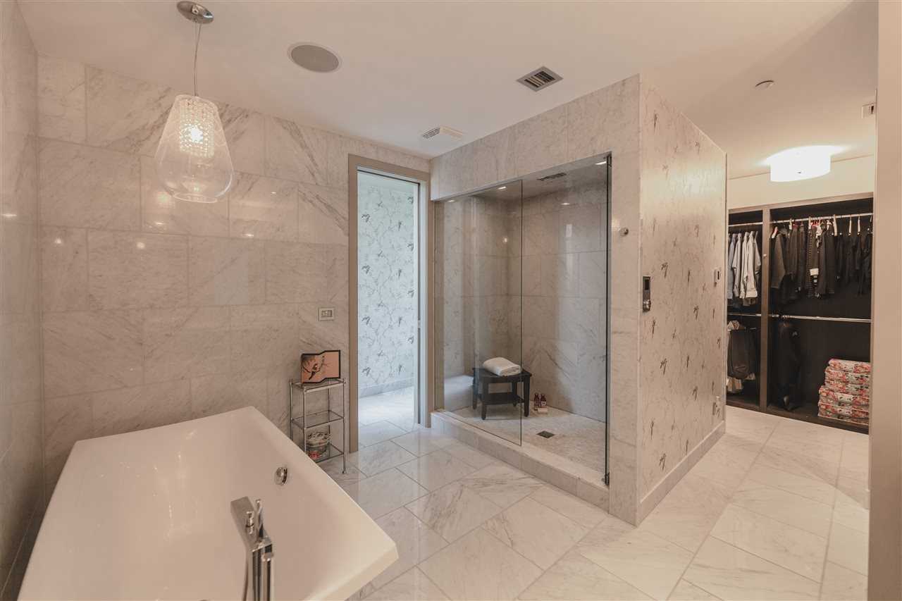 En suite with white tile floor and white marble walls, all speckled with black; freestanding soaker tub on the left, large shower with a black stool on the right; walk-through closet in background