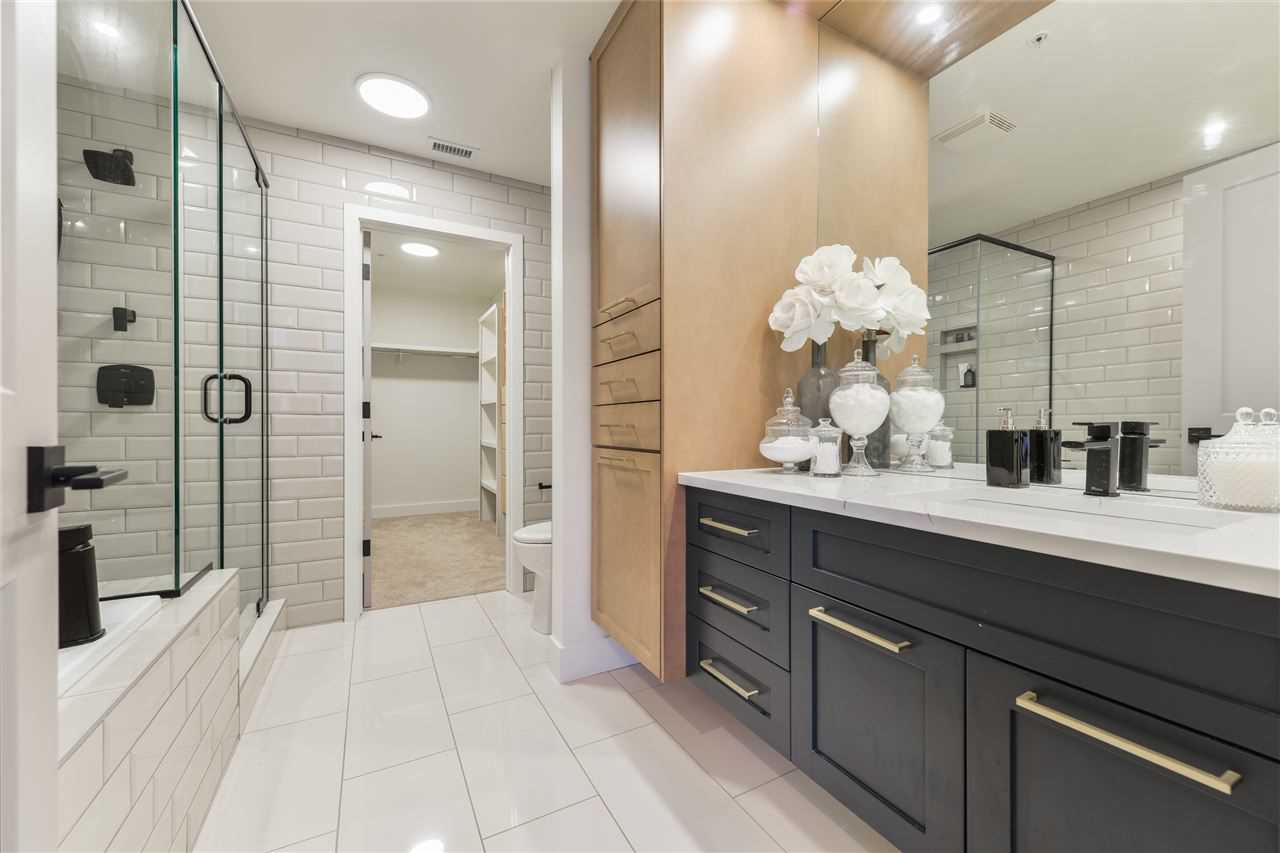 En suite bathroom with white walls and tile floor; black and beige cabinets, white countertop under large mirror; sit-down glass shower on the left