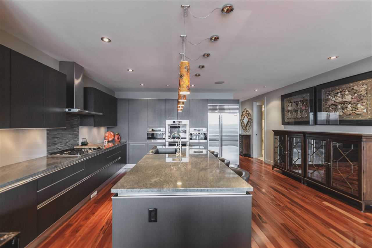 Interior kitchen with brown hardwood floor, black and grey cabinets, ceiling and walls; five yellow lights hanging over grey waterfall island; glass cabinets on the right