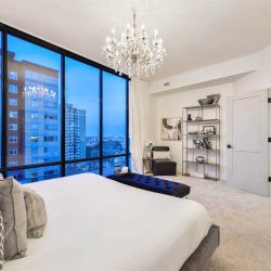 Master bedroom, white ceiling, walls, carpets and bed; hanging glass chandelier; floor-to-ceiling windows looking out to buildings on the left
