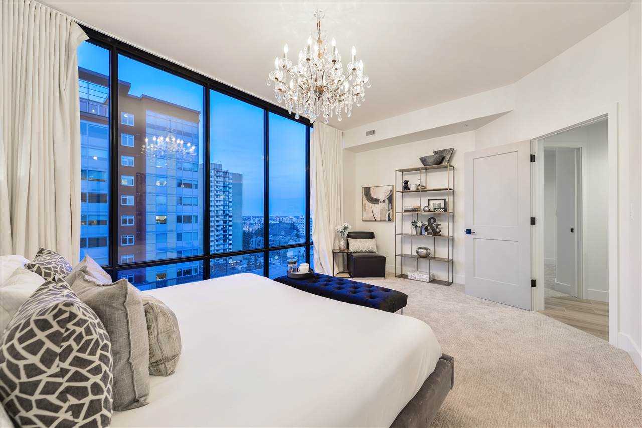 Master bedroom, white ceiling, walls, carpets and bed; hanging glass chandelier; floor-to-ceiling windows looking out to buildings on the left