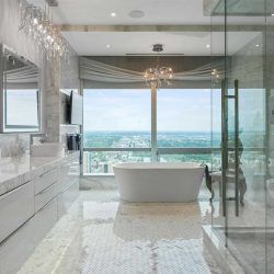 En suite with white tile floor, white cabinets, ceiling and walls, white marble countertop; glass shower on right; white freestanding soaker tub straight ahead, with large windows behind and chandelier above