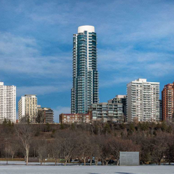 Exterior shot of circular condo tower, with blue windows and white roof, from a distance, in winter; leave-less trees in foreground; shorter white and brick condo buildings on either side