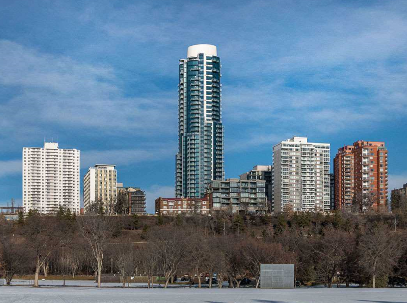 Exterior shot of circular condo tower, with blue windows and white roof, from a distance, in winter; leave-less trees in foreground; shorter white and brick condo buildings on either side
