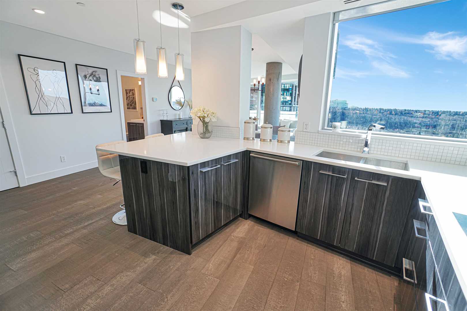 Interior condo kitchen, light hardwood floor, dark wood cabinets, white counters, ceiling and walls; large window by sink
